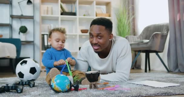 Simpático sonriente feliz cuidado de piel negra papá mirando a la cámara junto con su lindo hijo pequeño en casa, vista frontal, cámara lenta — Vídeo de stock