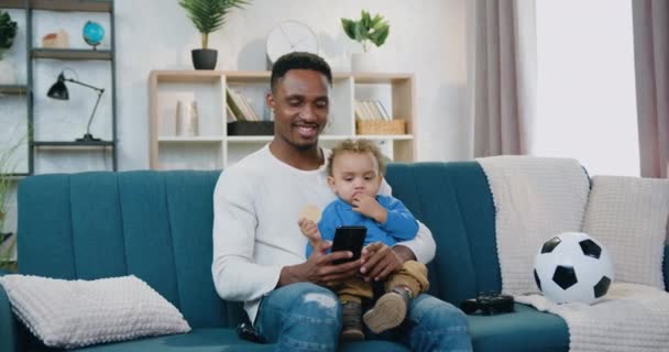 Портрет привабливого позитивного турботливого молодого чорношкірого батька, який тримає свого маленького сина на колінах і використовує свій мобільний, сидячи на м'якому дивані вдома — стокове відео