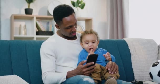 Close-up van sympathieke glimlachen gelukkig ontspannen liefdevolle donkere huid vader die het houden van zijn mooie kleine zoon op de knieën tijdens de beoordeling video 's op smartphone thuis — Stockvideo