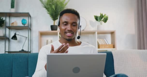 Retrato de atractivo sonriente confiado de alta cualificación de piel negra chico en los auriculares que la celebración de chat de vídeo en el ordenador, sentado en el sofá en casa — Vídeos de Stock