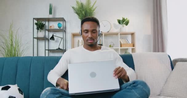 Przystojny pozytywny pewny siebie nowoczesny inteligentny czarny skórzany facet w słuchawki siedzi na kanapie w domu i zaczyna pracować na laptopie, widok z przodu — Wideo stockowe