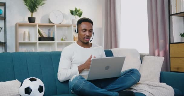 Schöne positive selbstbewusste freundliche junge Afroamerikanerin in Headset sitzt in entspannter Pose auf der Couch zu Hause beim Videochat mit Freunden am Computer — Stockvideo