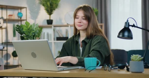 Attraktives, freudig zufriedenes junges Mädchen in lässiger Kleidung sitzt an ihrem Arbeitsplatz im Homeoffice und arbeitet mit traumhaftem Lächeln am Computer — Stockvideo