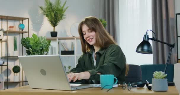 Retrato de una jovencita alegre y sonriente que se sienta frente a la computadora en casa y disfruta de la música favorita haciendo movimientos de baile — Vídeos de Stock