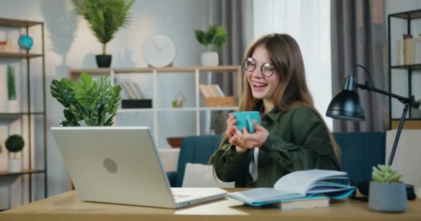 Encantadora y sonriente joven sin preocupaciones sosteniendo una taza de té mientras ve videos divertidos en la computadora sentada en su lugar de trabajo en la oficina en el hogar — Vídeos de Stock