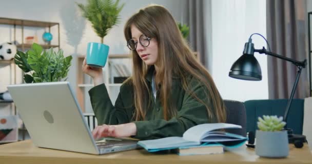魅力的な肯定的な自信を持ってスマート現代女性学生で眼鏡で彼女の職場で自宅に座って、コンピュータ上で大学の家庭の仕事で働く、お茶を飲む — ストック動画