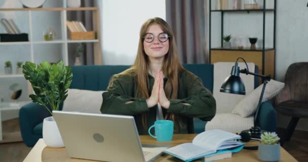 Close-up van goed uitziende glimlachende ontspannen jonge vrouw in een bril die thuis op haar werkplek zit tijdens het werken op de computer en mediteert met gesloten ogen om stilte en harmonie te voelen — Stockvideo