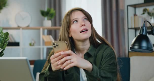 Verträumt positive schöne junge Mädchen in lässiger Kleidung sitzt an ihrem Arbeitsplatz zu Hause, wenn sie über Antwort denken, wenn sie mit Freunden in den sozialen Medien chatten — Stockvideo