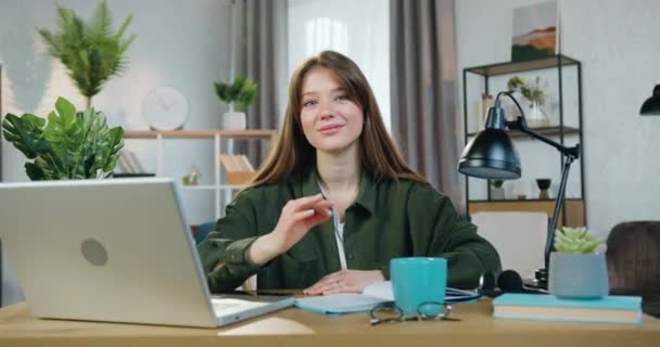Dość zadowolony zrelaksowany uśmiechnięty dziewczyna w casual ubrania siedzi przed kamerą w biurze w domu podczas pracy na laptopie i pokazując symbol ok, widok z przodu — Wideo stockowe