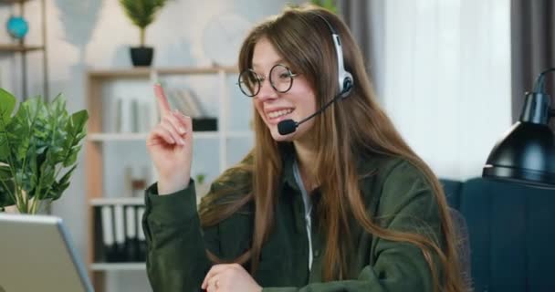 Довольно улыбаясь удовлетворены 25-летняя женщина фрилансер в наушниках, сидя перед компьютером во время онлайн-конференции с коллегами и давая им звуковые советы — стоковое видео