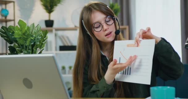 Bela confiante positivo 25-aged trabalhador do sexo feminino em óculos usa fone de ouvido discutindo relatório com gráfico durante videoconferência com colegas no laptop de casa — Vídeo de Stock