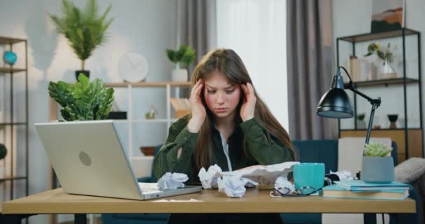 Перегруженная грустная 25-летняя женщина чувствует головную боль и массирует виски во время работы с финансовым отчетом, много скомканных бумаг лежит на столе — стоковое видео
