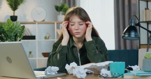 Крупным планом измученной молодой женщины с грустным лицом, сидящей перед камерой на своем рабочем месте со смятыми бумагами и массирующими висками — стоковое видео