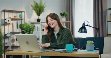 Çekici, gülümseyen, mutlu, zeki genç bir kız ev bilgisayarında çalışıyor ve cep telefonuyla konuşuyor aynı anda çay içiyor, ön tarafta, ağır çekimde.