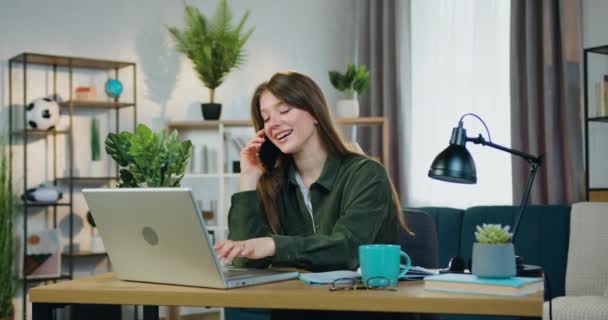 Привлекательная улыбающаяся счастливая молодая умная девушка, работающая за компьютером в домашнем офисе и разговаривающая на мобильном телефоне одновременно пьющая чай, вид спереди, замедленная съемка — стоковое видео