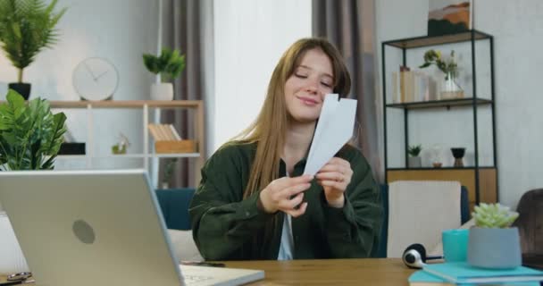 Портрет привлекательной улыбающейся счастливой беззаботной девушки с длинными прямыми волосами, которая запускает бумажный самолет, сидя на своем рабочем месте дома — стоковое видео