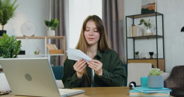 Schöne fröhliche junge braunhaarige Frau, die am Tisch im gemütlichen Homeoffice sitzt und Papierflugzeuge durch den Raum fliegen lässt, Frontansicht — Stockvideo