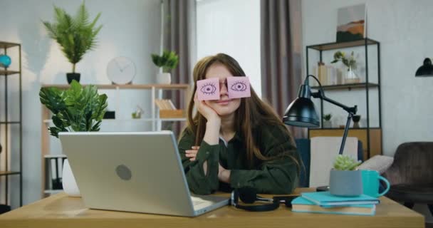 Retrato de atractiva hembra moderna divertida que se sienta en su lugar de trabajo en casa y posando en la cámara en gafas con pegatinas en las que dibujó los ojos abiertos — Vídeos de Stock