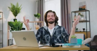 Çekici, güler yüzlü, rahat sakallı adam rahat bir odada masada oturuyor ve dizüstü bilgisayardaki işini durdururken gözleri kapalı meditasyon yapıyor.