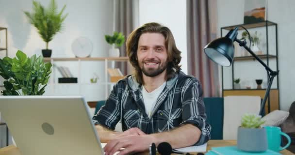 Portré jóképű mosolygós, magabiztos, fiatal szakállas férfi alkalmi ruhák, amelyek jelzik hüvelykujj fel a kamerába otthon iroda — Stock videók