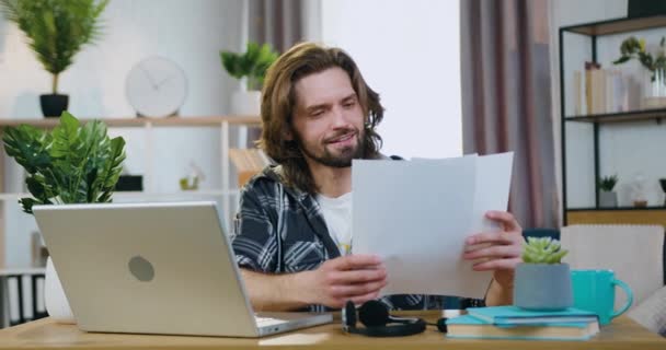 Όμορφος θετικός βέβαιος επιτυχημένος 30χρονος γενειοφόρος επιχειρηματίας απομακρυσμένος εργαζόμενος στο σπίτι με εταιρικά έγγραφα και αναλύοντας δεδομένα με πληροφορίες για το laptop — Αρχείο Βίντεο