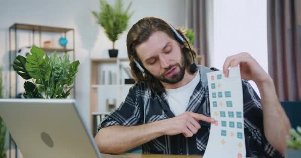 Probable confiado creativo joven barbudo oficinista en auriculares explicando informe financiero con estadísticas durante la videoconferencia en la computadora en la oficina en casa — Vídeos de Stock