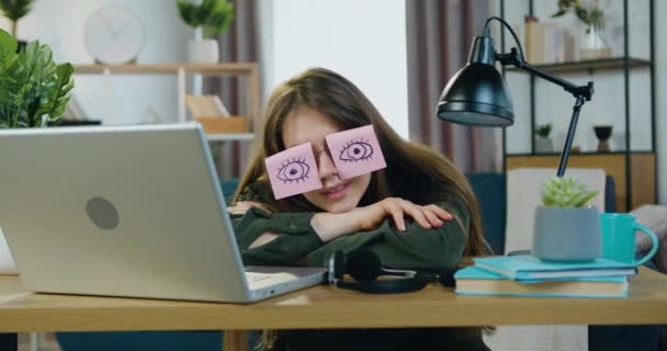 Красива втомлена сучасна молода жінка-працівник засинає на столі біля ноутбука і одягає окуляри з кумедними наклейками з намальованими відкритими очима, крупним планом — стокове відео