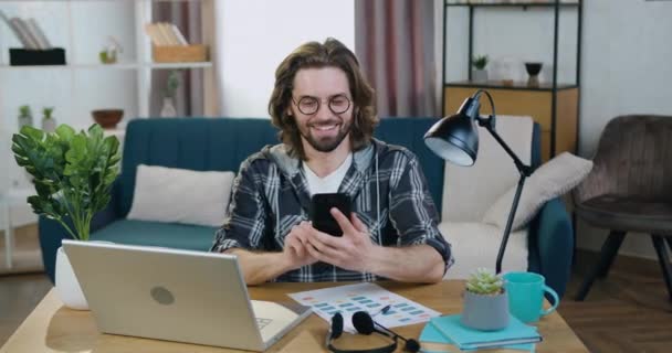 Aangename vrolijke glimlach moderne stijlvolle bebaarde man met bril zittend aan het bureau in gezellig thuiskantoor en met behulp van zijn smartphone bij het maken van pauze in zijn computer werk — Stockvideo