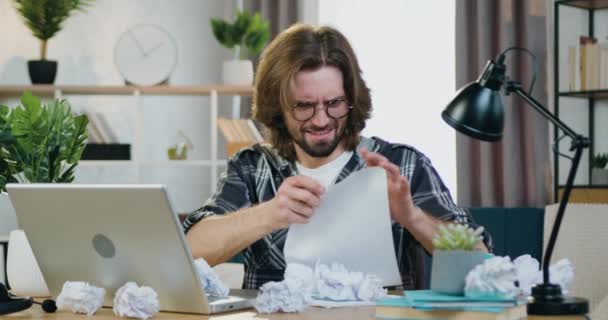 Frustré jeune employé de bureau barbu en colère dans des lunettes assis à son lieu de travail à la maison, travailler avec des papiers et insatisfait de l'information, les déchirer et jeter beahind son dos — Video