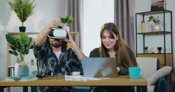 Guapo sonriente chico barbudo satisfecho usando auriculares de realidad virtual cerca de hermosa novia alegre que elegir visión futurista para él en el ordenador portátil en casa — Vídeo de stock