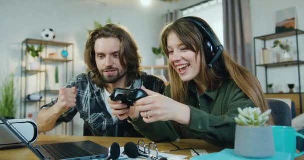 Закройте глаза на привлекательную улыбчивую возбужденную молодую пару, которая сидит перед компьютером дома и развлекается, когда девушка играет в игры с геймпадом — стоковое видео