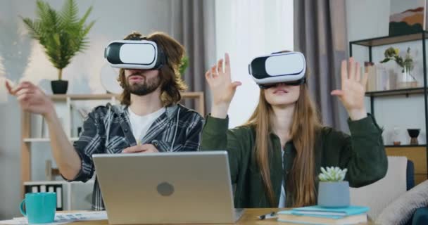 Roztomilý spokojený inteligentní mladý pár ve virtuální realitě sluchátka sedí na svém pracovišti v domácí kanceláři a pracuje na imaginární obrazovce, moderní technologie koncepce — Stock video