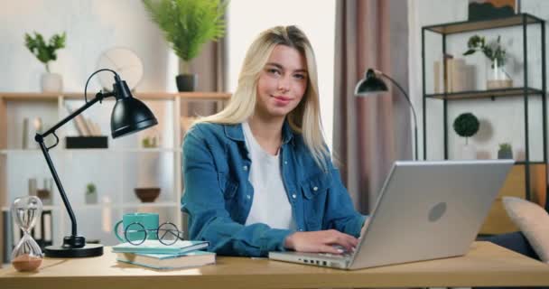 Gros plan de jolie jeune femme souriante de bonne humeur en chemise en jeans qui s'assoit à la table avec un ordinateur portable et regarde dans la caméra avec un visage satisfait — Video