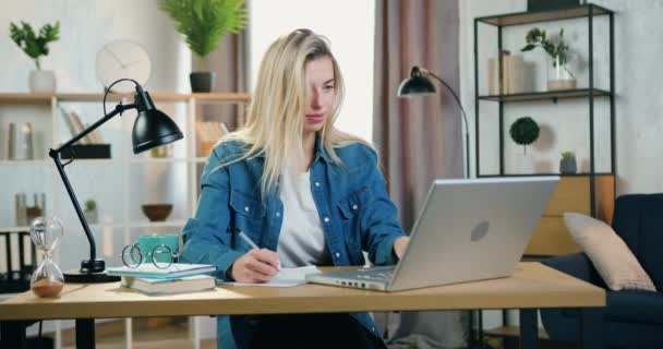 Distant work concept where good-looking positive confident blond young woman working with laptop and doing needed notes into papers in home office — Stock Video