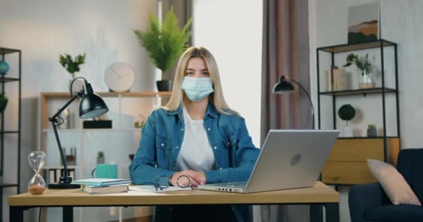 Концепція "Здоров'я та захист ковад-19", де добре виглядає серйозна молода блондинка в масці для обличчя, сидить перед камерою в затишному домашньому офісі — стокове відео