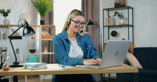 Blisko do przyjemnego uśmiechu zadowolona blondynka sukcesu freelancer w słuchawkach, które siedzi w miejscu pracy w domu i pracuje na laptopie, zwolnione tempo — Wideo stockowe