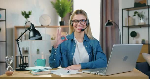 Zbliżenie z piękny radosny sukces młody blondynka-freelancer w bezprzewodowy zestaw słuchawkowy, który siedzi przed kamerą i pokazując symbol ok w przytulny dom biuro — Wideo stockowe