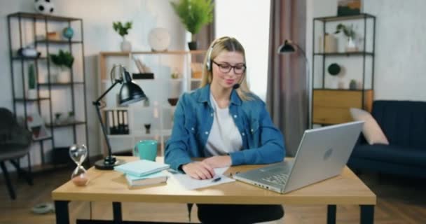 Câmera vista sobre sorrindo muito hábil jovem loira mulher em óculos e fones de ouvido que olhando para a câmera durante o trabalho no laptop em casa escritório — Vídeo de Stock