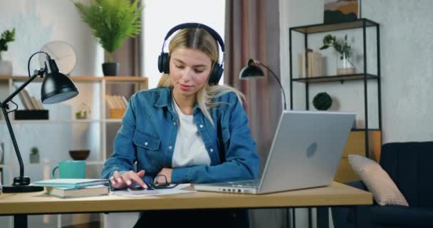 Portrait d'adorable jeune blonde souriante dans des écouteurs assis à son lieu de travail à la maison et appréciant les mélodies préférées de la playlist téléphonique — Video