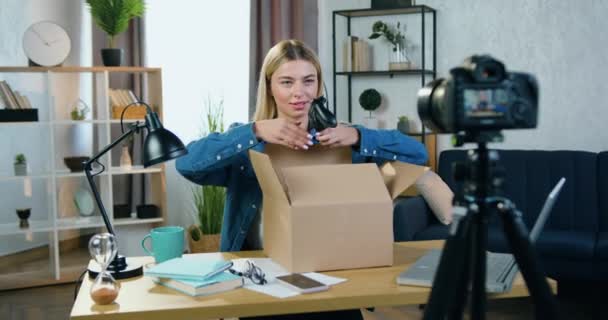 Хорошо выглядящая счастливая блондинка-блондинка, снимающая процесс распаковки в домашней студии и показывающая новый геймпад в камеру — стоковое видео