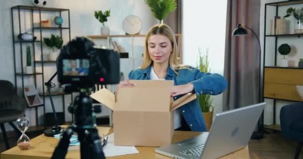 Приваблива щаслива здивована молода блондинка записує свою реакцію при розпакуванні картонної коробки на камеру в домашній студії — стокове відео