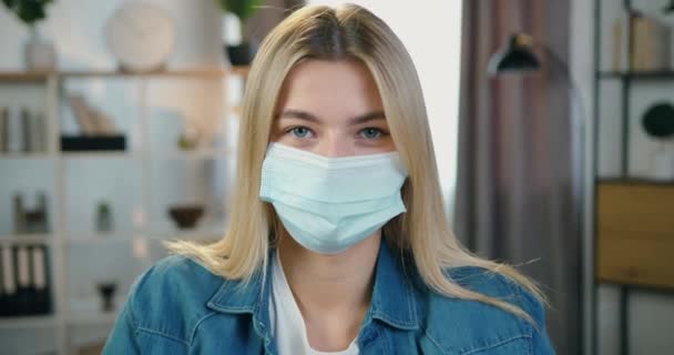 Zbliżenie kobiecego portretu twarzy w masce ochronnej podczas samoizolacji w czasach pandemii koronawirusowej na tle współczesnego pokoju w domu — Wideo stockowe