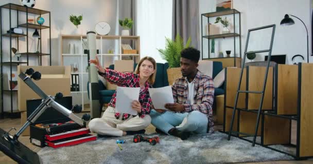 Atrakcyjny szczęśliwy uśmiechnięty mieszany wyścig para siedzi razem na podłodze we własnym domu i omawia projektowanie wnętrz za pomocą kreatywnych pomysłów napisanych na papierach — Wideo stockowe
