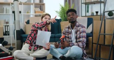 Oda tasarımı konsepti. Sevimli, genç, çok ırklı bir çift kendi evinde yerde oturuyor ve kağıt üzerinde tasvir edilmiş planlarına uygun mobilya yapma fikirlerini tartışıyor.