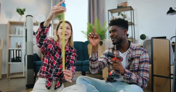 Aantrekkelijke glimlachende moderne multiraciale paar samen zitten op de vloer in hun nieuwe appartement en bespreken kamer interier ontwerp gezien de grootte van bepaalde items van meubilair — Stockvideo