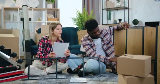 Goed uitziende vrolijke glimlachende gemengde ras paar zitten op het tapijt in hun nieuwe huis en het meten van de afmetingen van planken met behulp van tape maatregel — Stockvideo