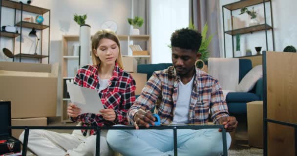 De cerca de adorable feliz sonriente joven pareja multiétnica que se sienta tanto en el suelo, chica viendo cómo su novio de piel negra medir la construcción de metal con cinta métrica — Vídeos de Stock