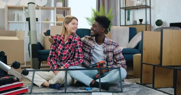 Portret van schattige vrolijke lachende jonge gemengde ras paar dat knuffelen in hun nieuw gekochte huis en kijken in de camera op de achtergrond van nog niet volledig uitgeruste kamer — Stockvideo