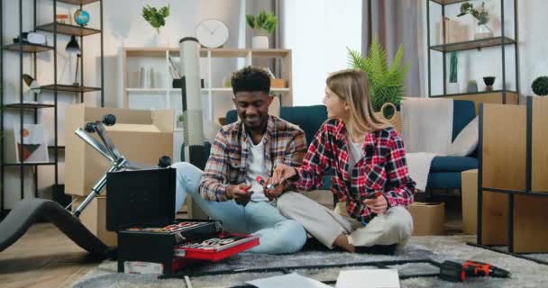 Atrakcyjny pozytywny nowoczesny młody wielorasowy para siedzi na podłodze w ich nowym mieszkaniu i próbuje wybrać niepotrzebny rozmiar wiertarki do ręcznego śrubokręta — Wideo stockowe