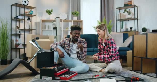 Close up on good-looking funny happy smiling mixed race couple which sitting on the floor in new bought house and playing with wrenches like swords between assembling the chair — ストック動画